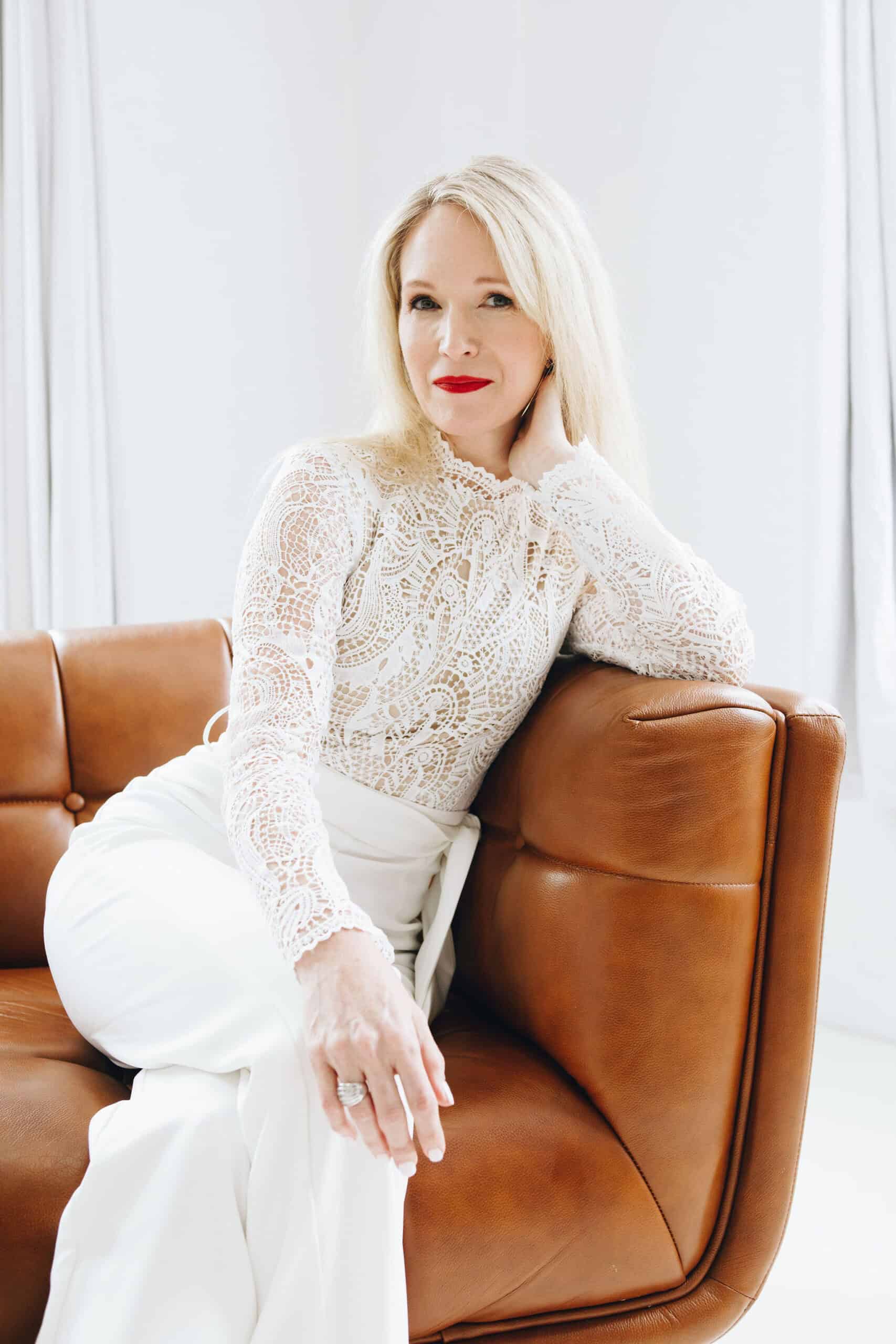 Beautiful woman in white lace top and white pants sitting on a tan leather couch in a bright room.