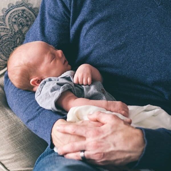 A man in a blue sweater gently cradles a sleeping newborn baby in his arms. The baby is dressed in a grey outfit and wrapped in a white blanket.