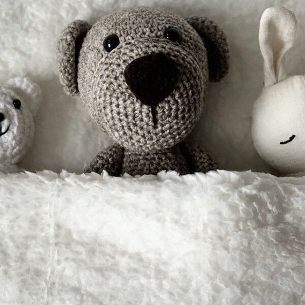 Three stuffed animals are tucked under a soft, white blanket. In the center is a crocheted bear with a brown nose and black eyes.