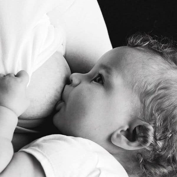 A baby with curly hair is breastfeeding, latched onto the mother's breast. The baby is looking up, and the scene captures a tender and intimate moment between mother and child.